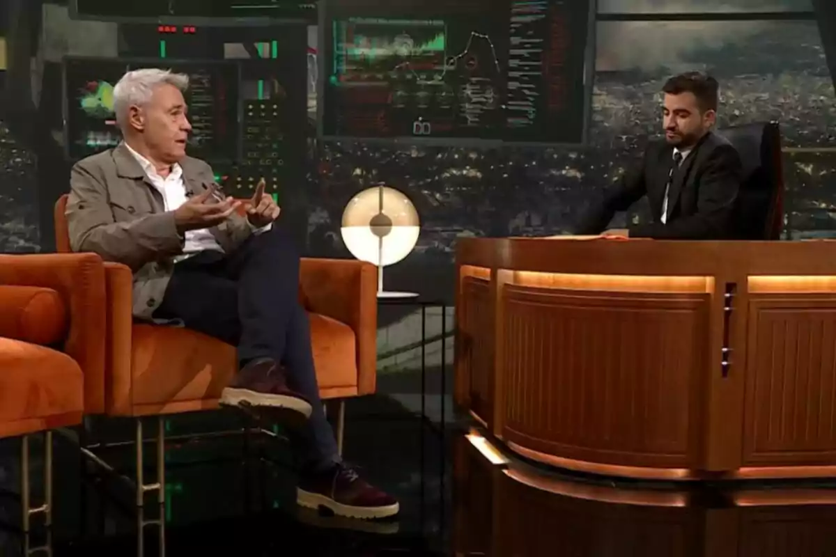Jordi González y Ricard Ustrell conversan en un set de televisión de Col·lapse uno sentado en un sillón naranja y el otro detrás de un escritorio de madera con pantallas y una lámpara de fondo Jordi González y Ricard Ustrell conversan en un set de televisión de Col·lapse uno sentado en un sillón naranja y el otro detrás de un escritorio de madera con pantallas y una lámpara de fondo