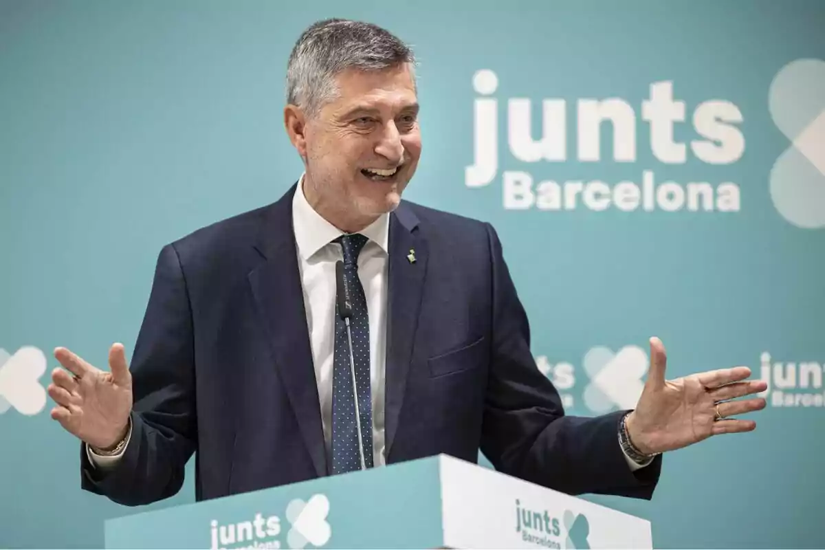 Un hombre en traje sonríe mientras habla en un podio con el logotipo de Un hombre en traje sonríe mientras habla en un podio con el logotipo de