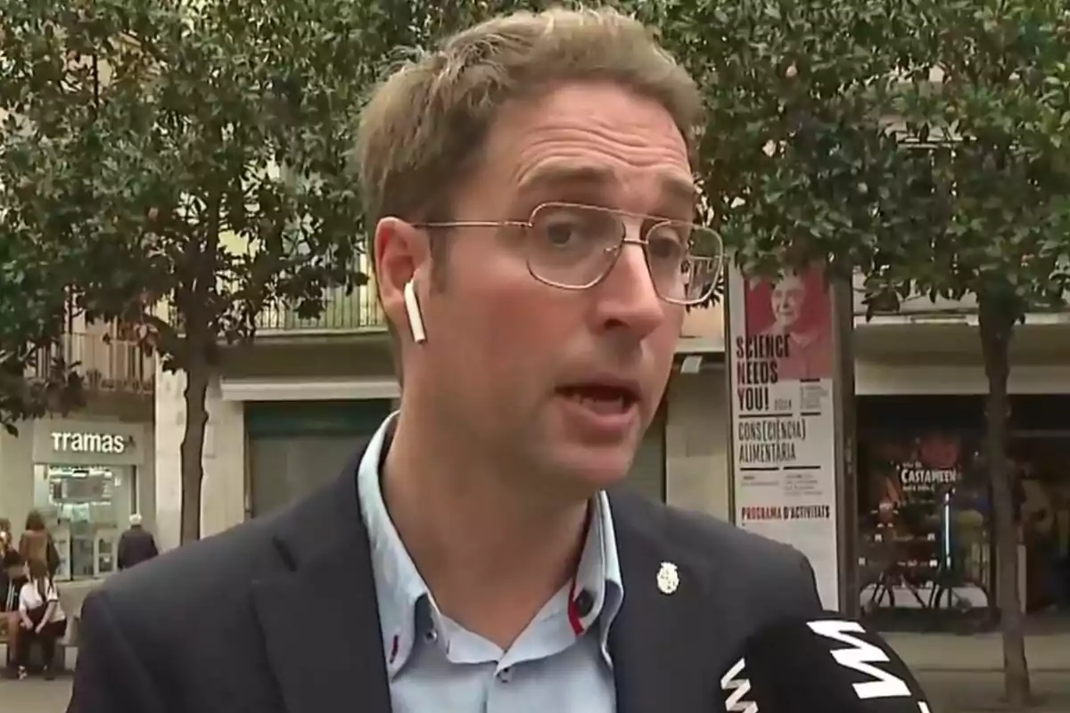 Un hombre con gafas y auriculares está hablando frente a un micrófono en una calle con árboles y carteles al fondo.