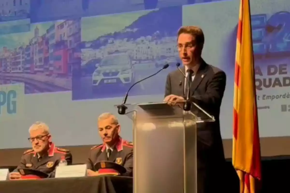Un hombre en un podio habla en un evento oficial junto a dos oficiales uniformados sentados, con una bandera a la derecha y un fondo con imágenes de una ciudad y un coche.