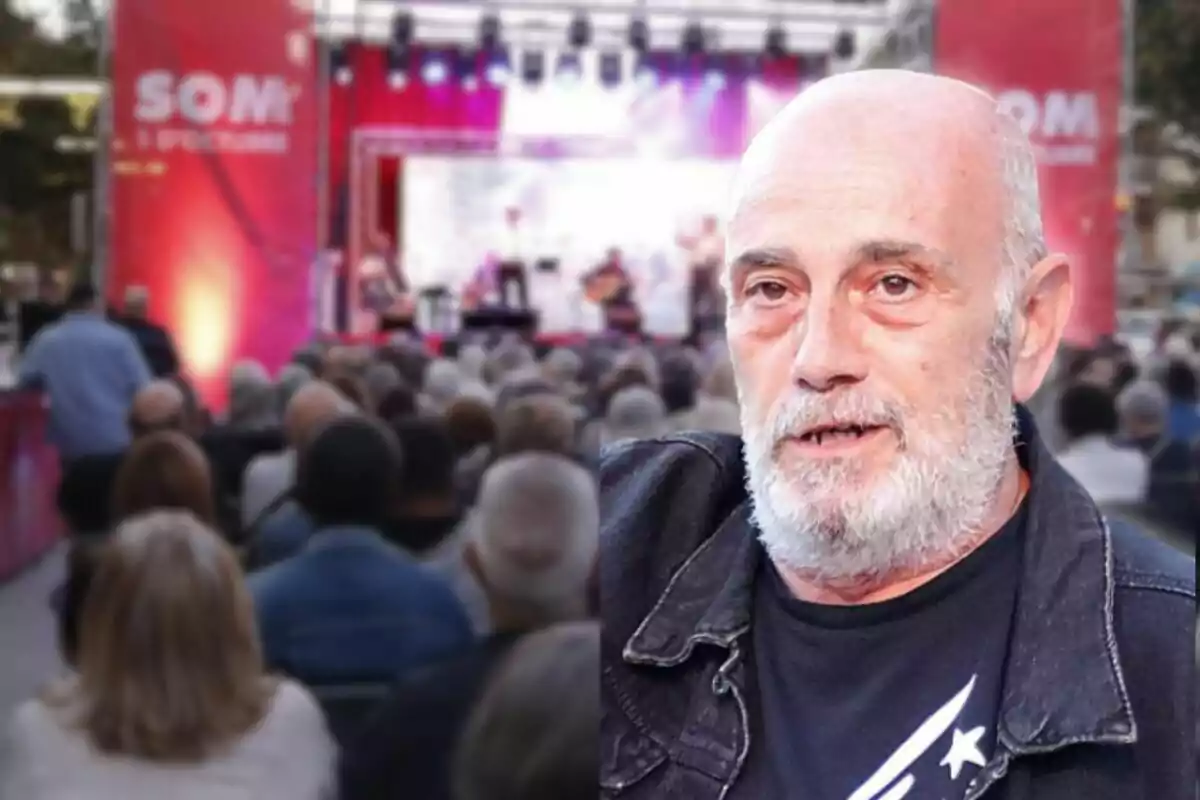 Un hombre de barba blanca en primer plano con un fondo de un evento al aire libre con un escenario iluminado y una multitud de personas.