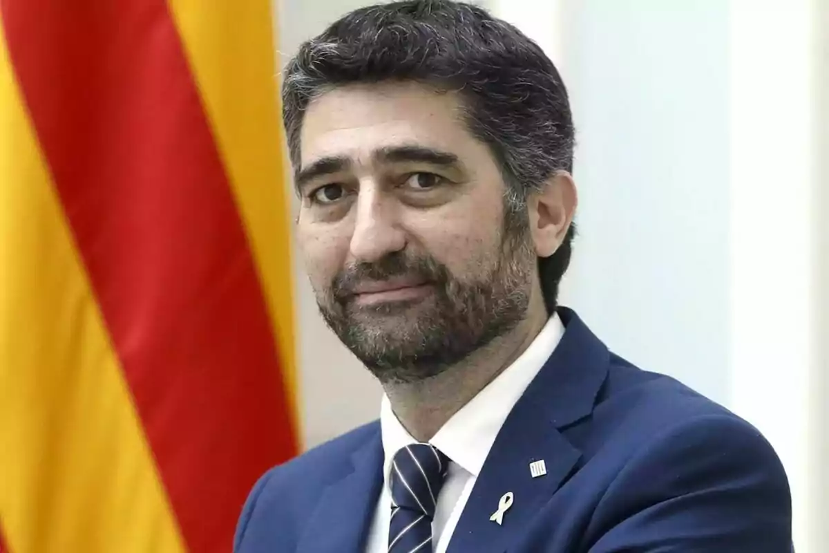 Un hombre con traje azul y corbata a rayas posa frente a una bandera con franjas rojas y amarillas.