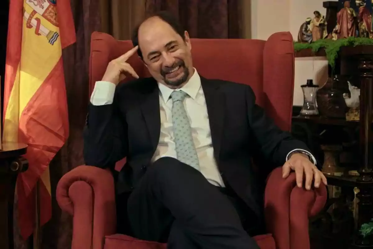 Jordi Sánchez como Antonio Recio sonriente con traje y corbata está sentado en un sillón rojo, con una bandera de España a su lado y un belén en el fondo en La que se avecina.