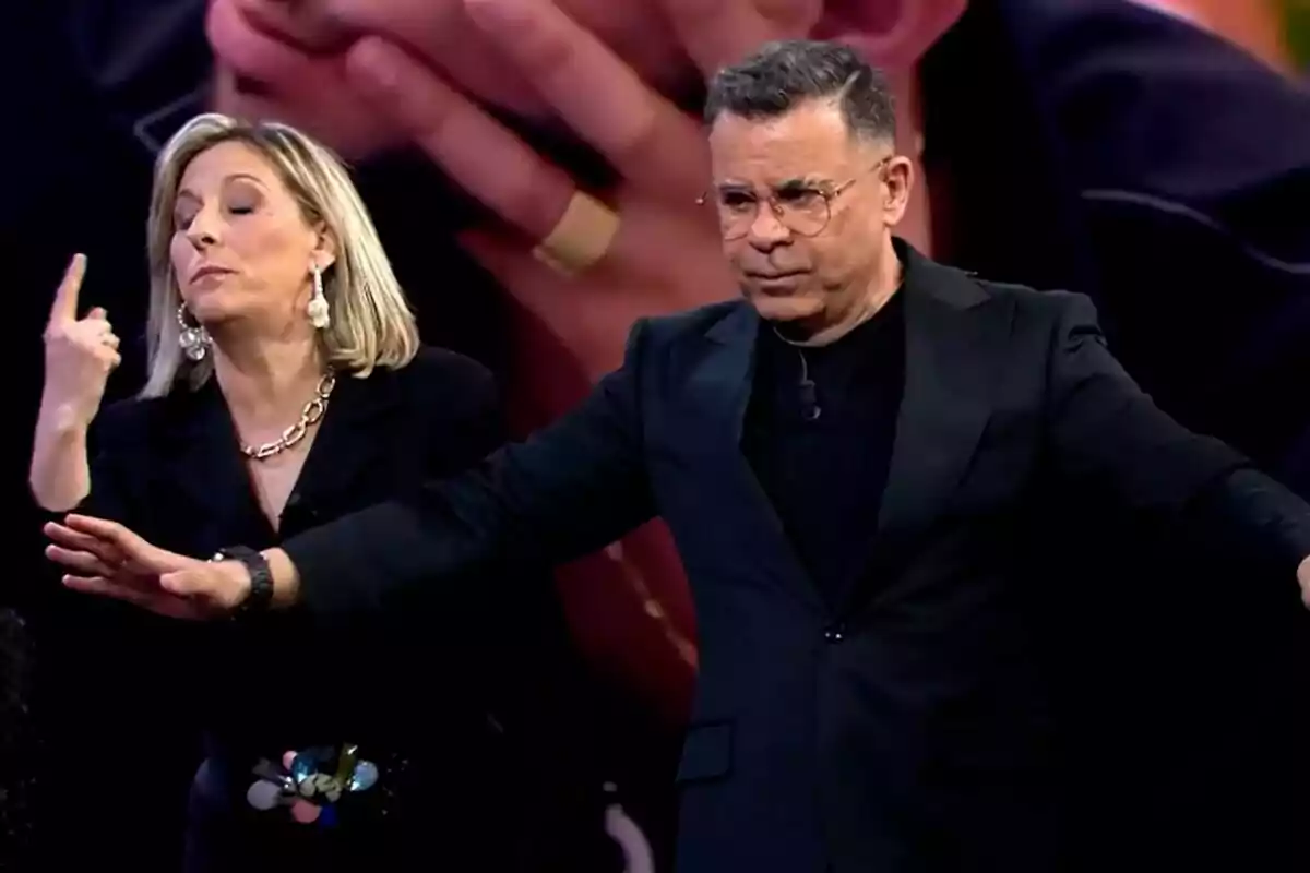 Dos personas en el plató de Gran Hermano, la madre de Violeta con gesto expresivo y Jorge Javier Vázquez con gafas y traje oscuro, ambos parecen estar en medio de una conversación tensa.