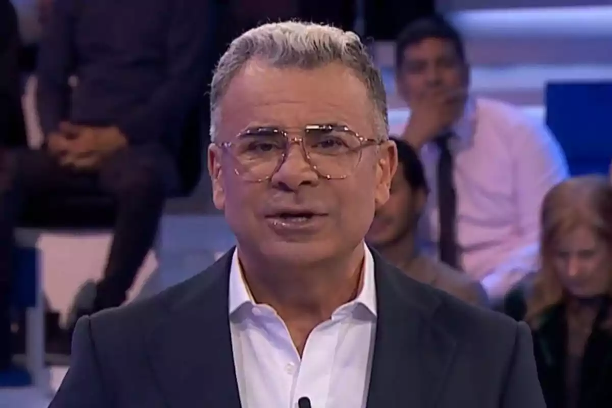 Jorge Javier Vázquez con gafas y cabello canoso está hablando en un estudio de televisión con personas sentadas al fondo en Hay una cosa que te quiero decir.