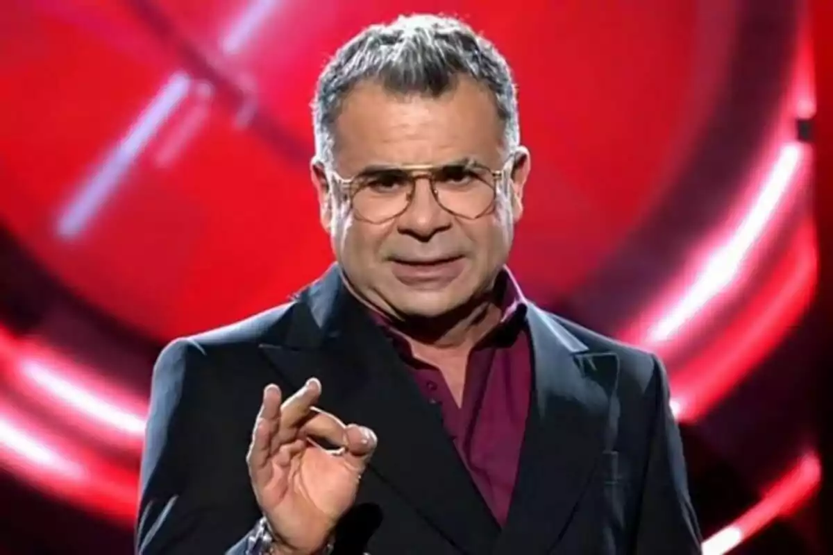 Jorge Javier Vázquez con gafas y traje oscuro gesticula con la mano frente a un fondo rojo en el plató de Gran Hermano.