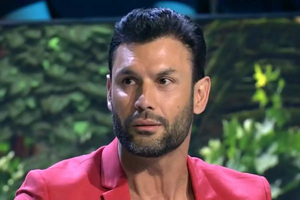 Jorge Pérez con barba y cabello oscuro lleva una chaqueta roja, con un fondo de vegetación desenfocada en Supervivientes.