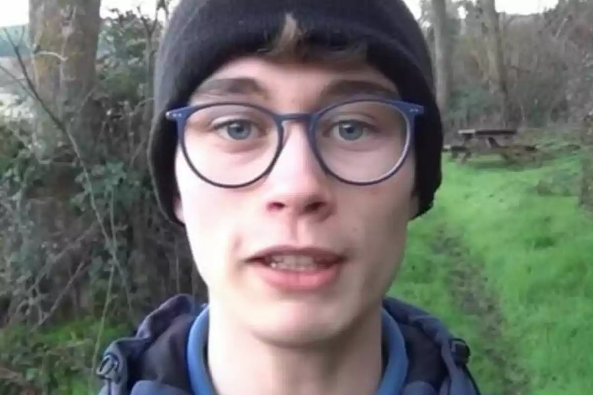 Un joven con gafas y gorro negro en un entorno natural con árboles y césped.