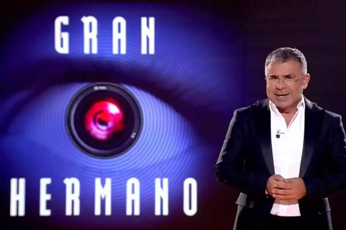 Jorge Javier Vázquez con un traje oscuro y camisa blanca, al lado de un logotipo que dice "Gran Hermano" con un ojo y una cámara en el centro.