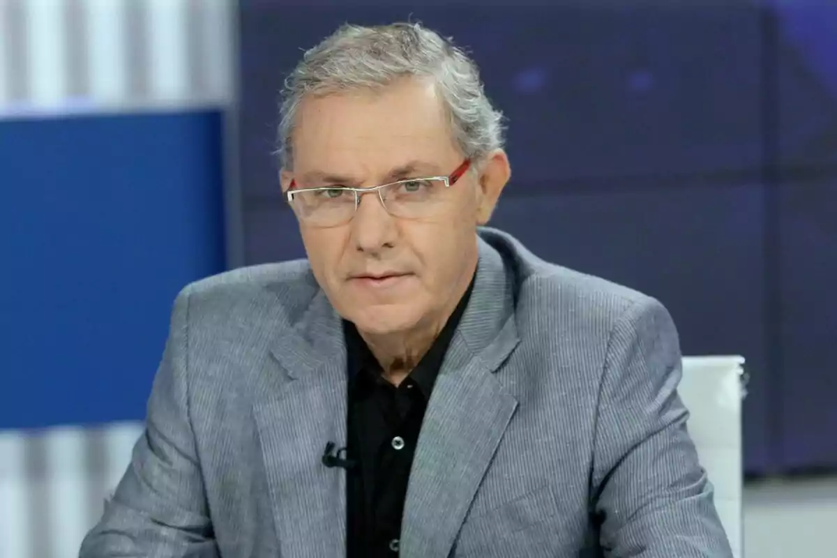 José Ángel de la Casa con gafas y cabello canoso lleva un traje gris y camisa negra mientras está sentado en un estudio de televisión. José Ángel de la Casa con gafas y cabello canoso lleva un traje gris y camisa negra mientras está sentado en un estudio de televisión.