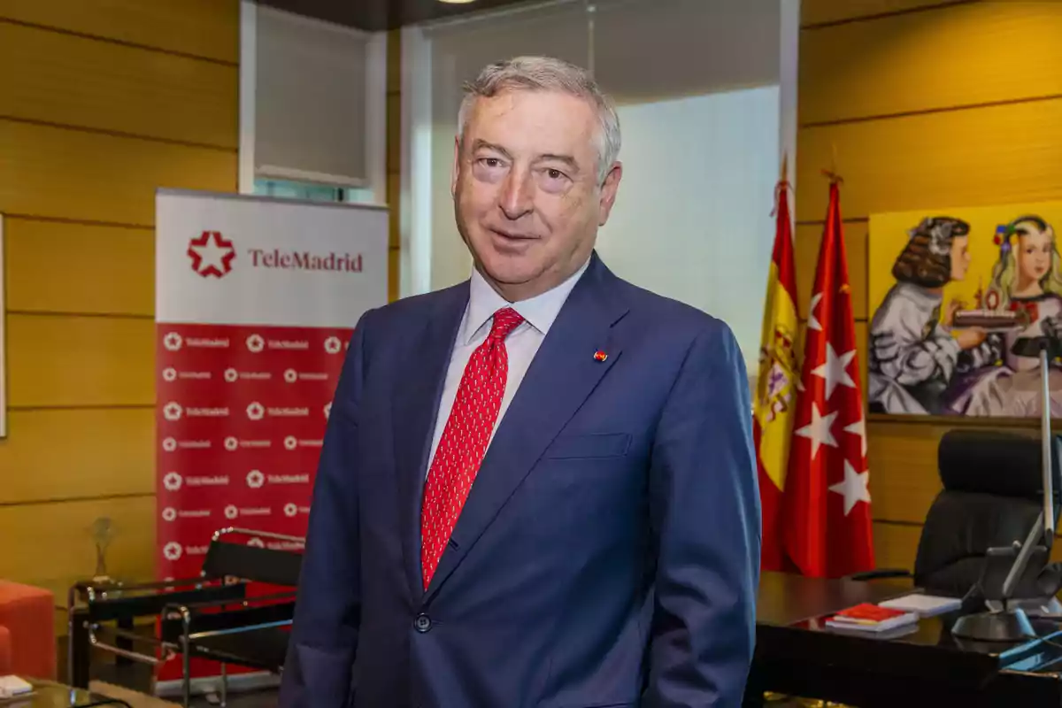 Fotografía de José Antonio Sánchez de traje azul y corbata roja en una oficina con banderas y un cartel de Telemadrid en el fondo.