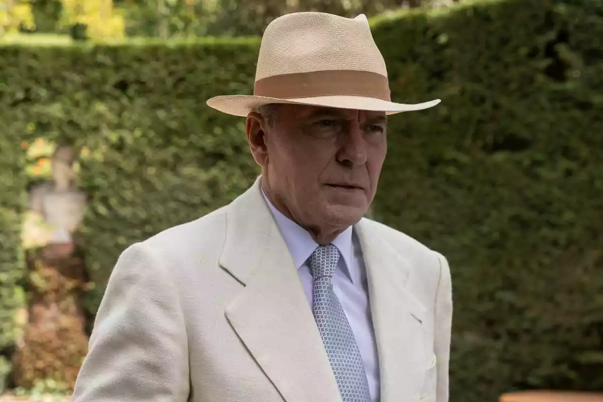 José Coronado con sombrero de paja y traje claro en un jardín en El Problema Final.