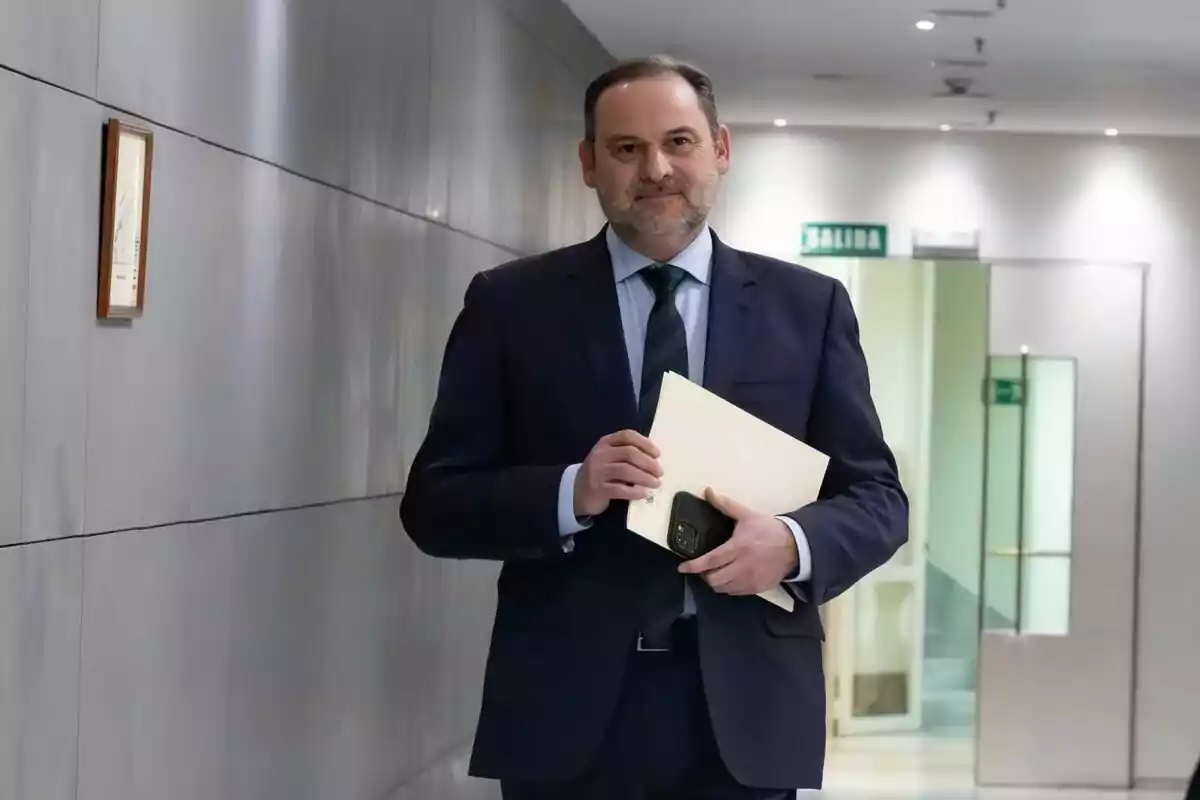 José Luis Ábalos con traje oscuro sostiene un sobre y un teléfono mientras camina por un pasillo iluminado. José Luis Ábalos con traje oscuro sostiene un sobre y un teléfono mientras camina por un pasillo iluminado.