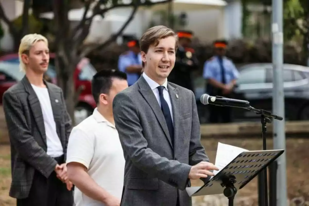Un hombre joven con traje gris habla en un micrófono al aire libre mientras otras personas están de pie detrás de él. Un hombre joven con traje gris habla en un micrófono al aire libre mientras otras personas están de pie detrás de él.