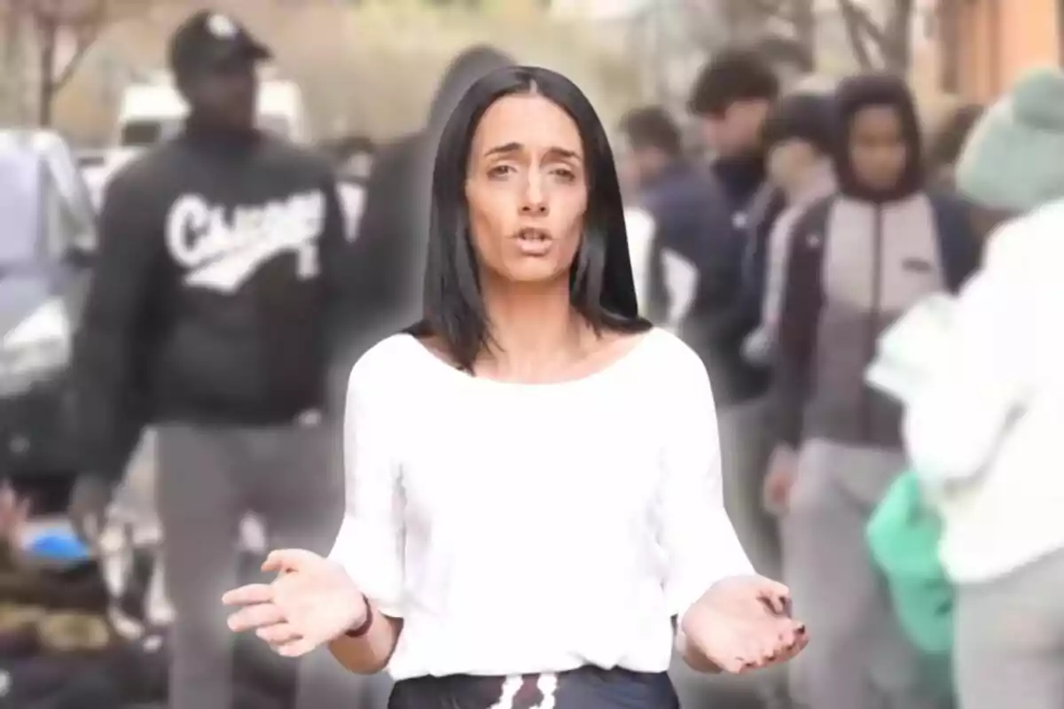 Una mujer de cabello oscuro y blusa blanca está hablando frente a un grupo de personas desenfocadas en el fondo.