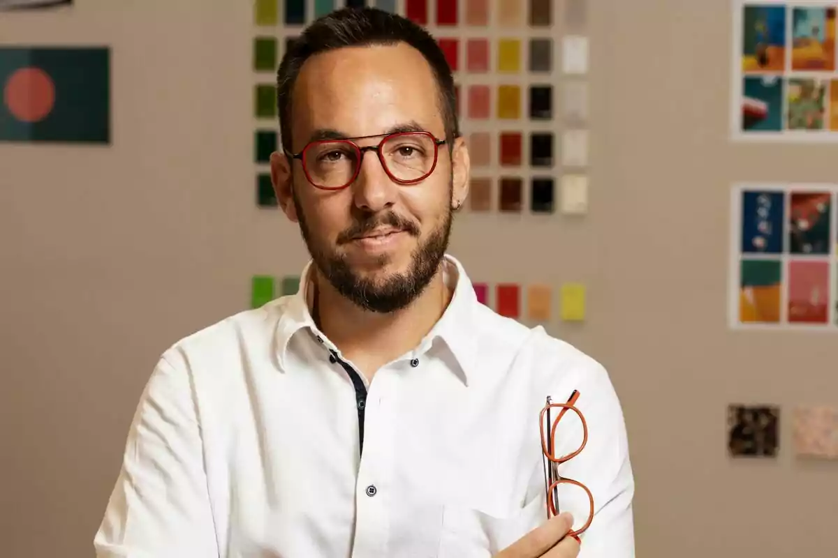 Josep Dosta, CEO de Woodys Eyewear, con gafas rojas y camisa blanca sostiene un par de gafas en una mano frente a una pared con muestras de colores. Josep Dosta, CEO de Woodys Eyewear, con gafas rojas y camisa blanca sostiene un par de gafas en una mano frente a una pared con muestras de colores.
