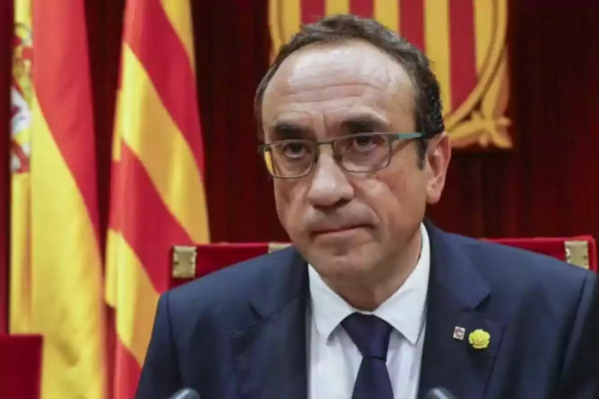 Un hombre con gafas y traje oscuro está sentado frente a banderas con franjas rojas y amarillas.