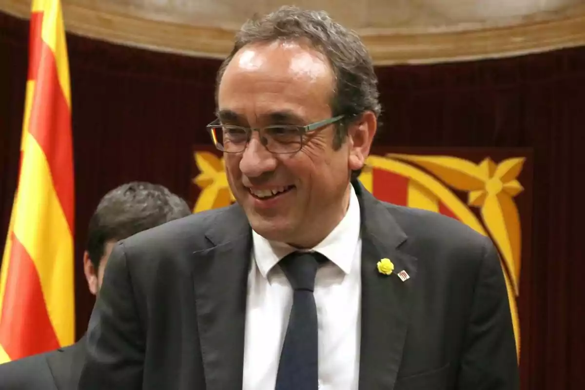 Josep Rull con gafas y traje oscuro sonriendo frente a una bandera catalana.