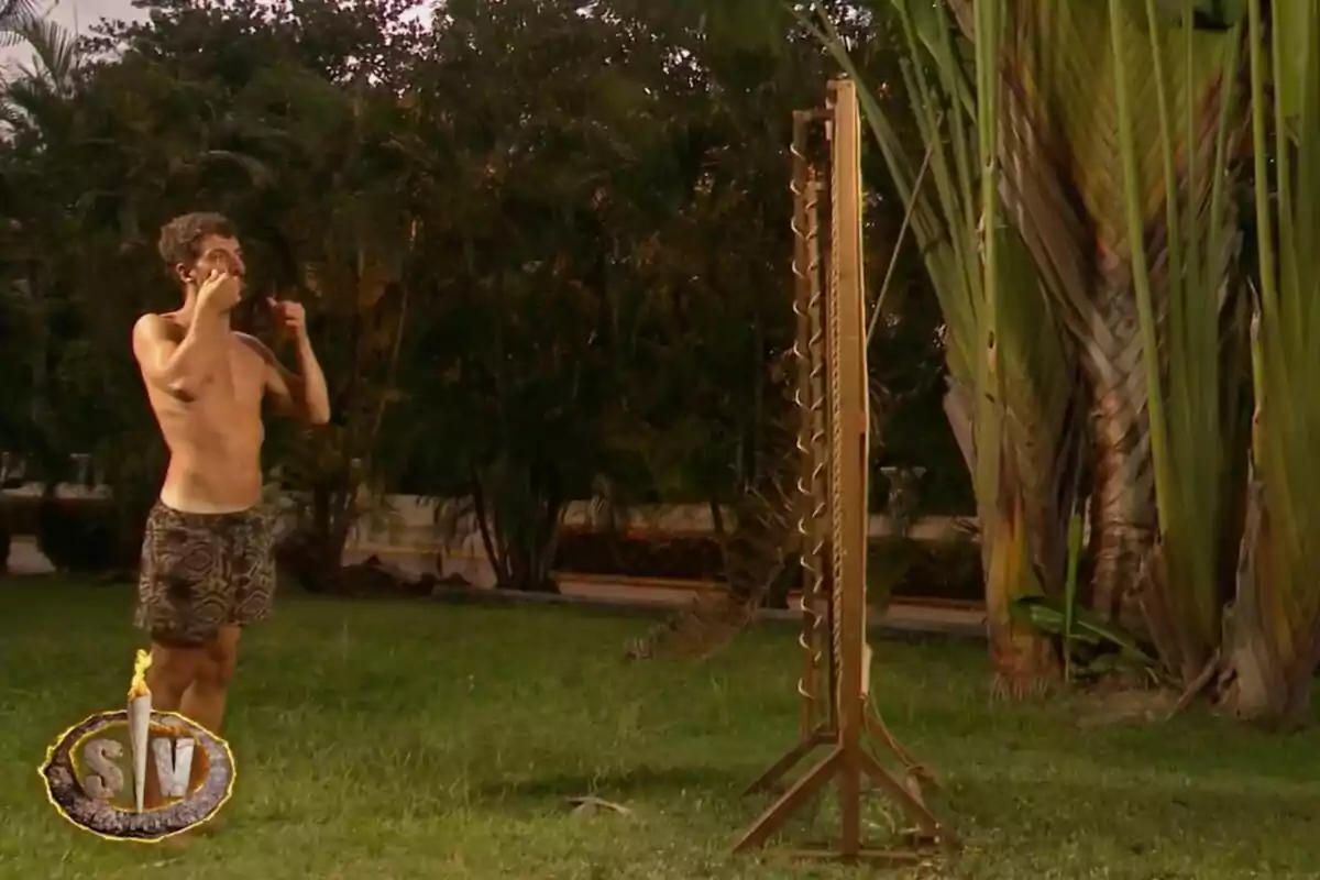Joshua Velázquez sin camiseta con pantalones cortos de pie en el césped frente a una estructura vertical de madera con cuerdas en un entorno al aire libre rodeado de vegetación en 'Supervivientes'. Joshua Velázquez sin camiseta con pantalones cortos de pie en el césped frente a una estructura vertical de madera con cuerdas en un entorno al aire libre rodeado de vegetación en 'Supervivientes'.