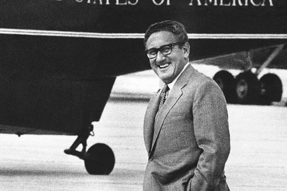Henry Kissinger sonriendo a la cámara con un avión de EE.UU. detrás