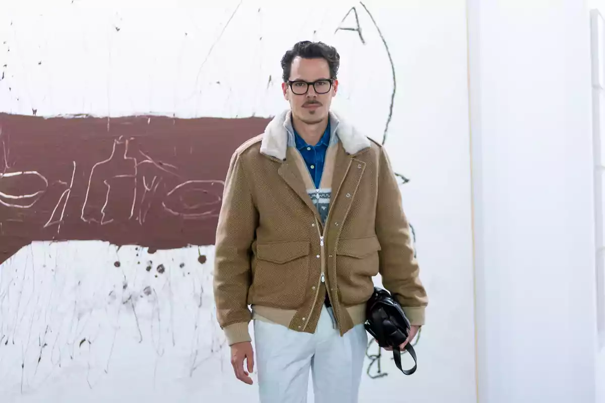 Juan Avellaneda con gafas y bigote lleva una chaqueta marrón con cuello de lana y sostiene un bolso negro, posando frente a una obra de arte abstracta en una galería. Juan Avellaneda con gafas y bigote lleva una chaqueta marrón con cuello de lana y sostiene un bolso negro, posando frente a una obra de arte abstracta en una galería.