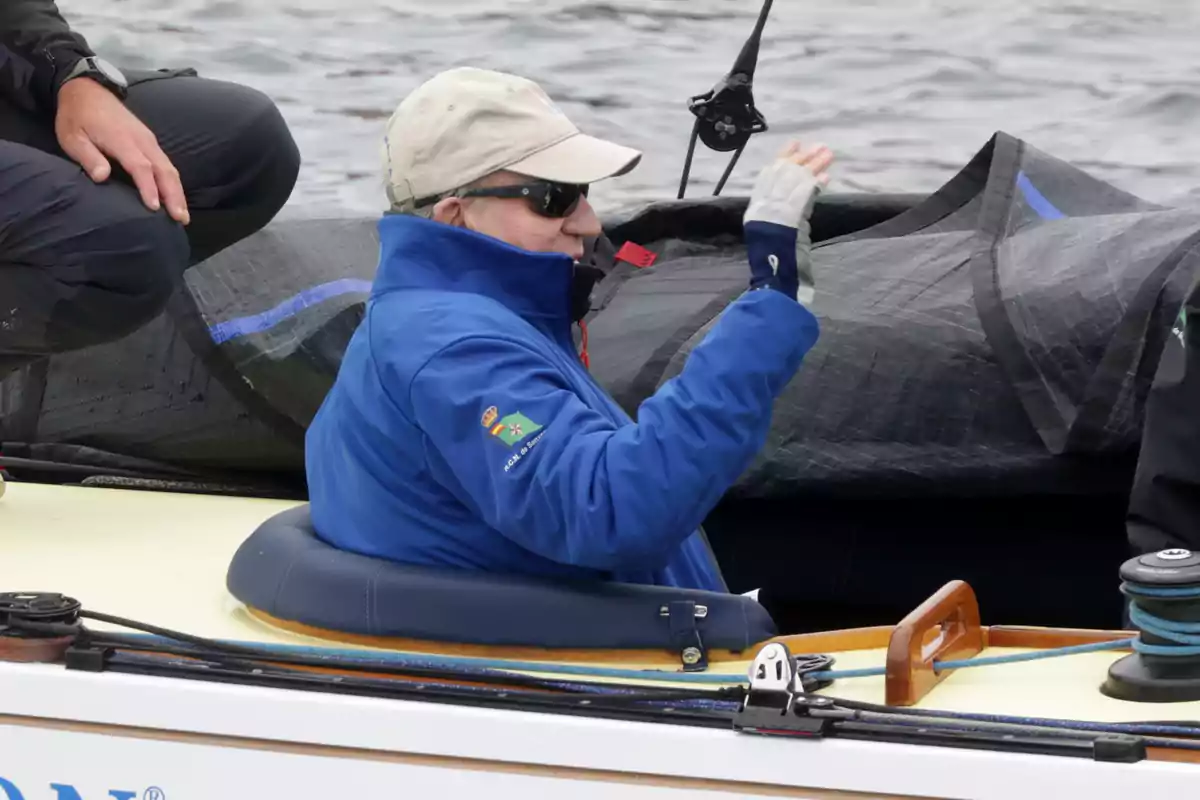 Juan Carlos I con chaqueta azul y gorra beige está sentado en un bote, saludando mientras lleva gafas de sol. Juan Carlos I con chaqueta azul y gorra beige está sentado en un bote, saludando mientras lleva gafas de sol.