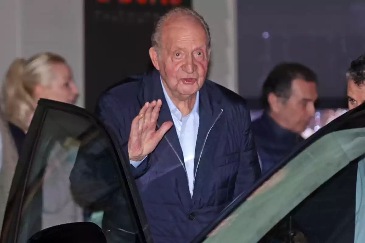 Juan Carlos I con chaqueta azul saluda mientras se encuentra junto a un automóvil, con otras personas en el fondo. Juan Carlos I con chaqueta azul saluda mientras se encuentra junto a un automóvil, con otras personas en el fondo.