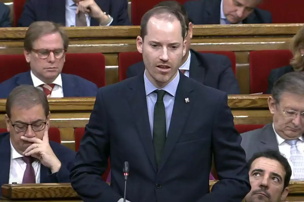 Un hombre de traje oscuro habla en un parlamento rodeado de otras personas sentadas.