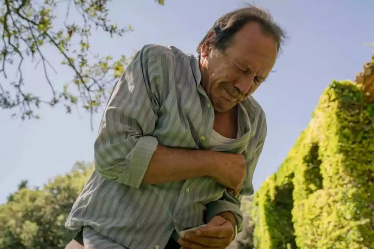 Juan Gea con expresión de dolor sujetándose el pecho en un jardín soleado en Sueños de Libertad.