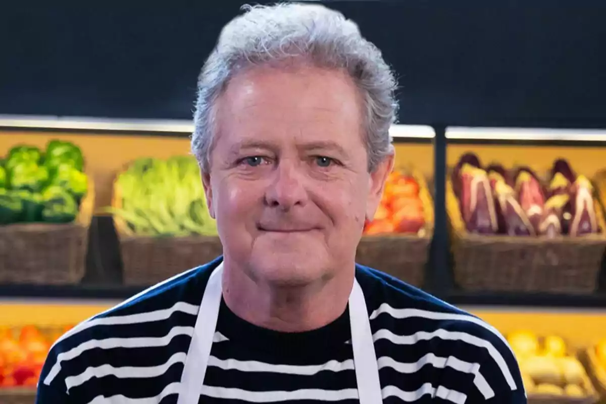 Juan Luis Cano con cabello canoso y suéter a rayas, usando un delantal blanco, con fondo de vegetales en un mercado en MasterChef Celebrity.