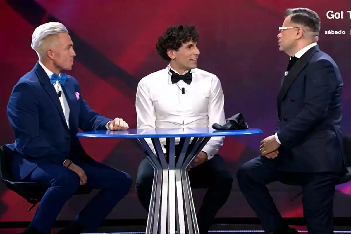 Juan, Óscar y Jorge Javier Vázquez vestidos formalmente conversan alrededor de una mesa azul en Gran Hermano con fondo rojo.