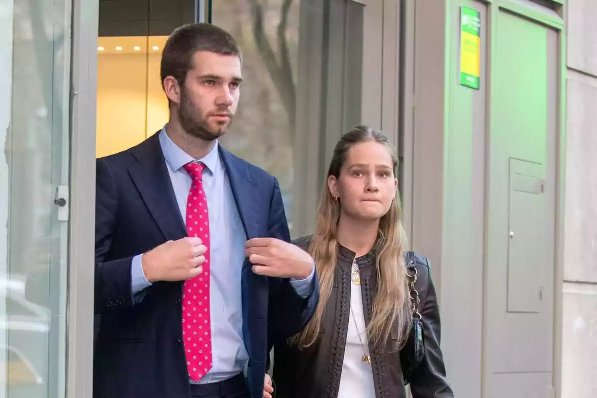 Juan Urdangarin serio en traje azul colocándoselo y corbata roja junto a Irene Urdangarin con los labios apretados y cazadora