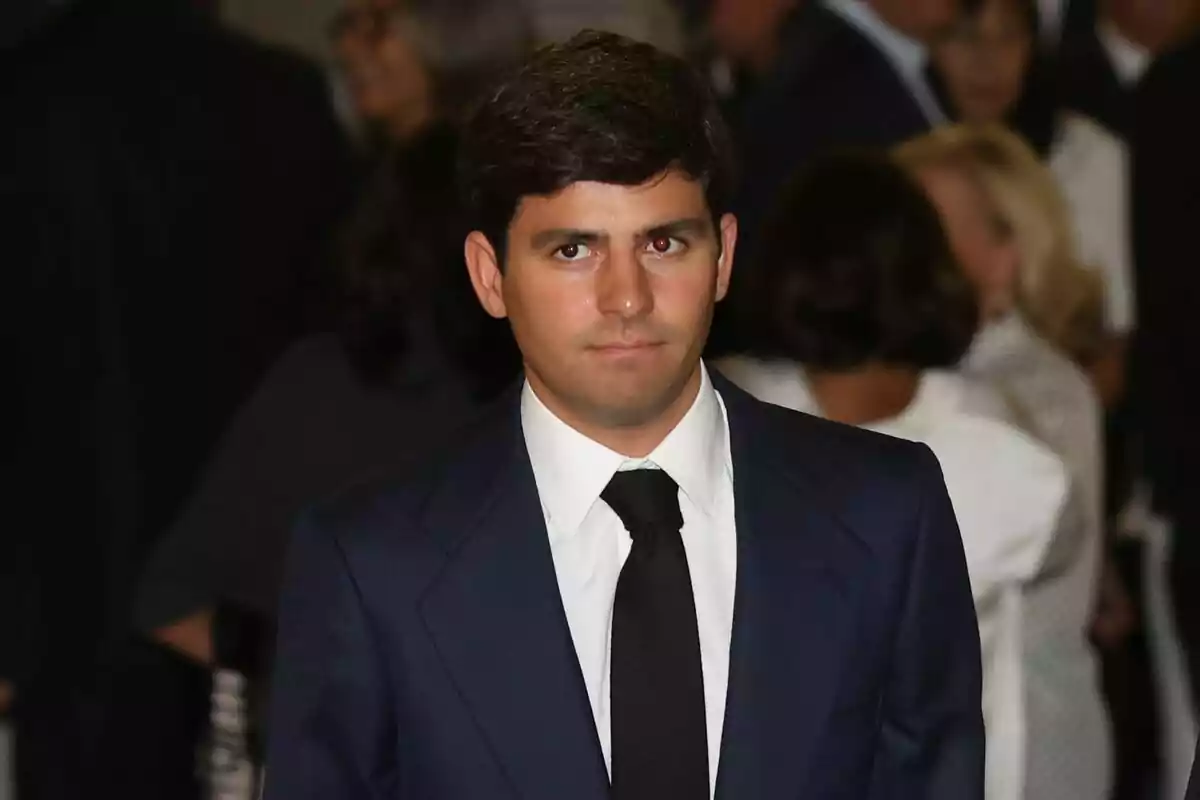 Plano corto de Juan Urquijo con cabello oscuro y corto, vestido con un traje azul marino, camisa blanca y corbata negra, está de pie en un entorno con varias personas en el fondo. Plano corto de Juan Urquijo con cabello oscuro y corto, vestido con un traje azul marino, camisa blanca y corbata negra, está de pie en un entorno con varias personas en el fondo.