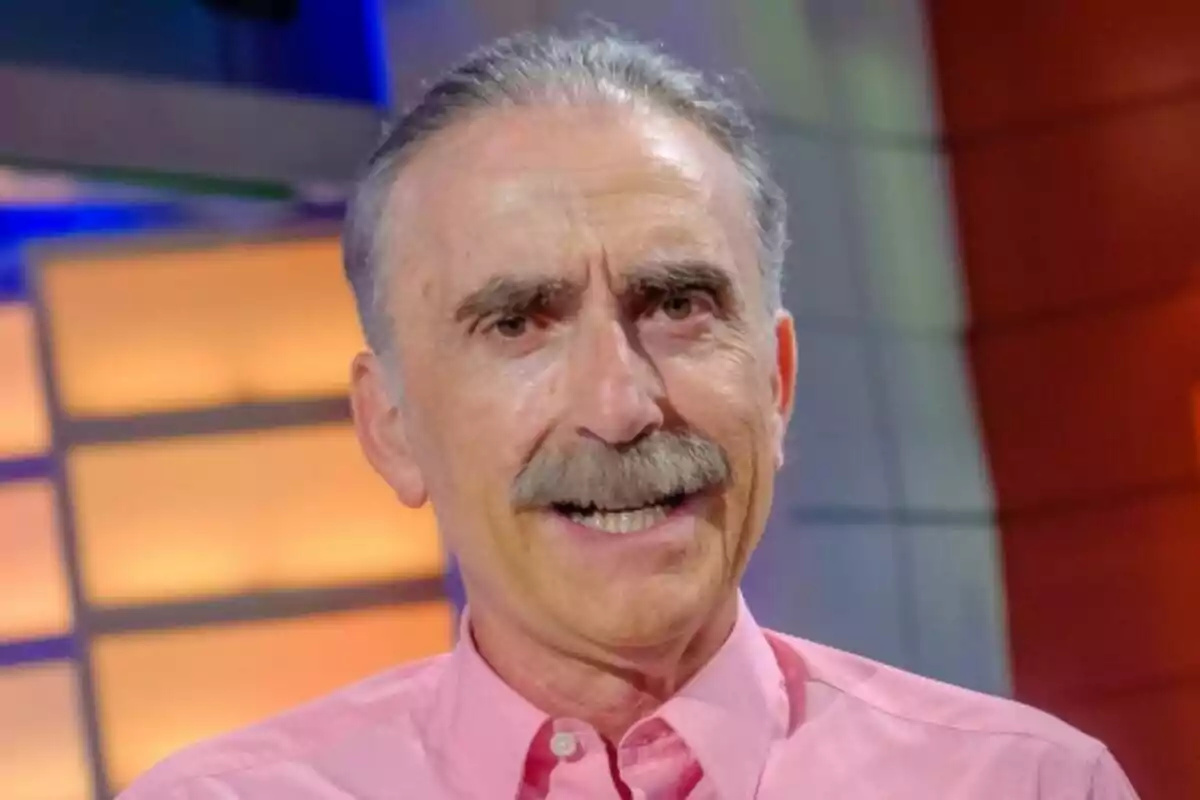 Juan y Medio de cabello canoso y bigote sonríe mientras lleva una camisa rosa en un entorno con luces cálidas en La tarde, aquí y ahora.