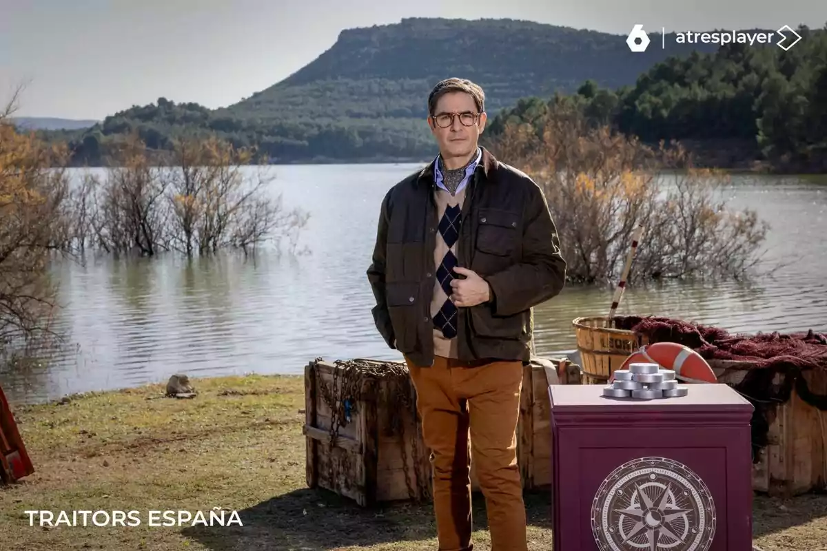 Juanra Bonet de pie junto a un lago con colinas y árboles al fondo, rodeado de cajas de madera y una mesa con un símbolo en Traitors España.