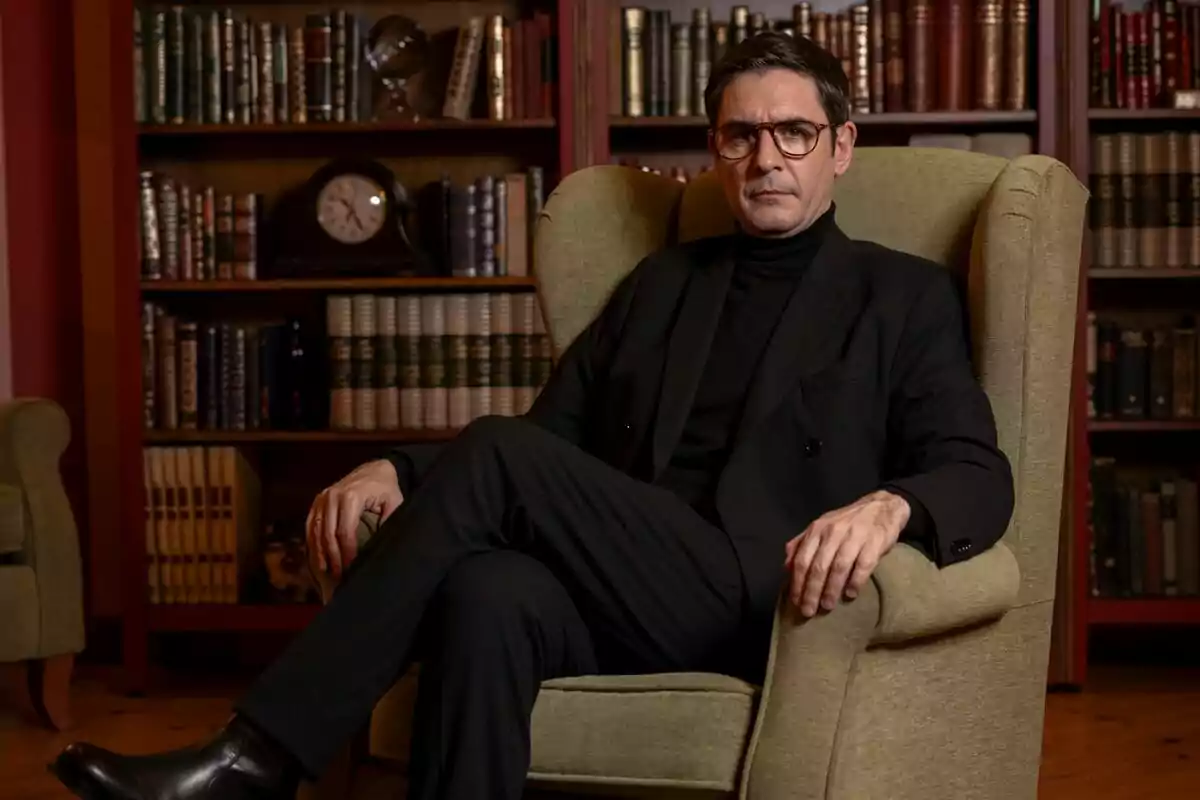 Juanra Bonet con gafas y traje negro está sentado en un sillón verde en una biblioteca con estanterías llenas de libros y un reloj en Traitors. Juanra Bonet con gafas y traje negro está sentado en un sillón verde en una biblioteca con estanterías llenas de libros y un reloj en Traitors.