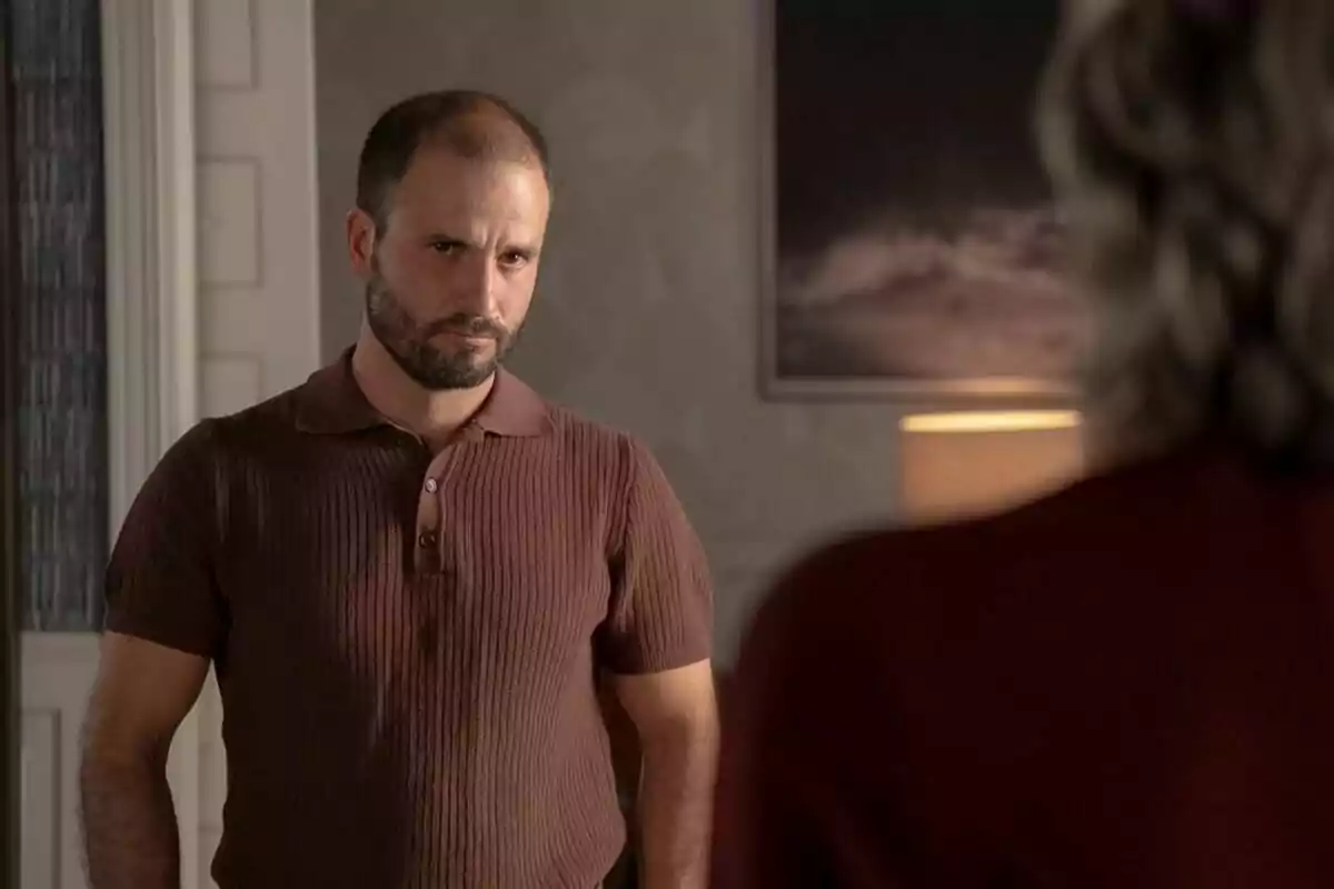 Julián Ortega con barba y camiseta marrón está mirando seriamente a otra persona en una habitación iluminada con una lámpara y un cuadro en la pared en la serie 4 estrellas.