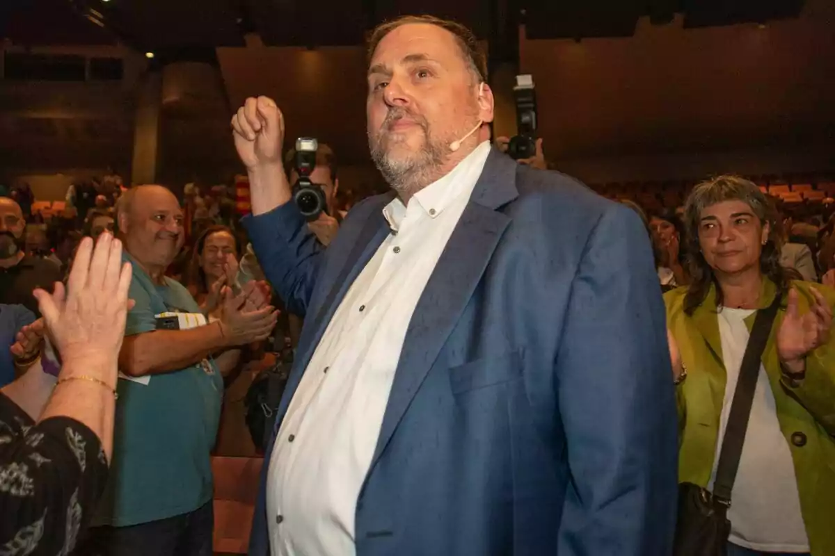 Oriol Junqueras con traje azul y camisa blanca levanta el brazo mientras es aplaudido por una multitud en un evento, con fotógrafos capturando el momento.