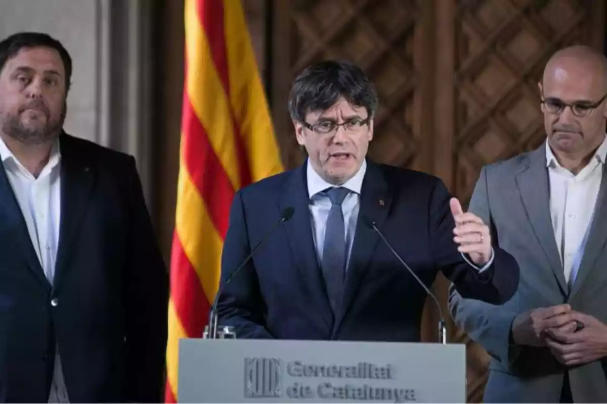 plano medio de Puigdemont hablando delante de dos micrófonos, detrás de él hay Junqueras y Romeva serios escuchándole