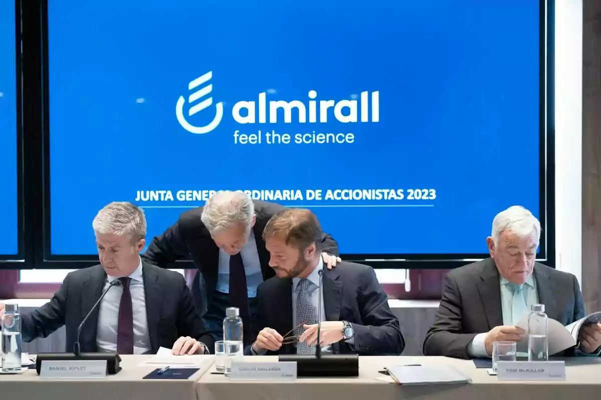 Reunión de la Junta General Ordinaria de Accionistas 2023 de Almirall, con varios hombres sentados en una mesa frente a un gran monitor azul con el logotipo de la empresa y el lema Reunión de la Junta General Ordinaria de Accionistas 2023 de Almirall, con varios hombres sentados en una mesa frente a un gran monitor azul con el logotipo de la empresa y el lema