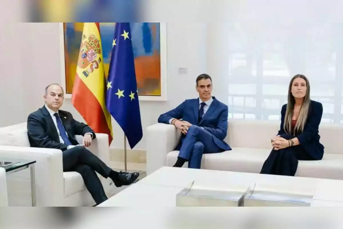 Tres personas con vestimenta formal sentadas en sofás blancos en una sala de reuniones con las banderas de España y la Unión Europea al fondo Tres personas con vestimenta formal sentadas en sofás blancos en una sala de reuniones con las banderas de España y la Unión Europea al fondo