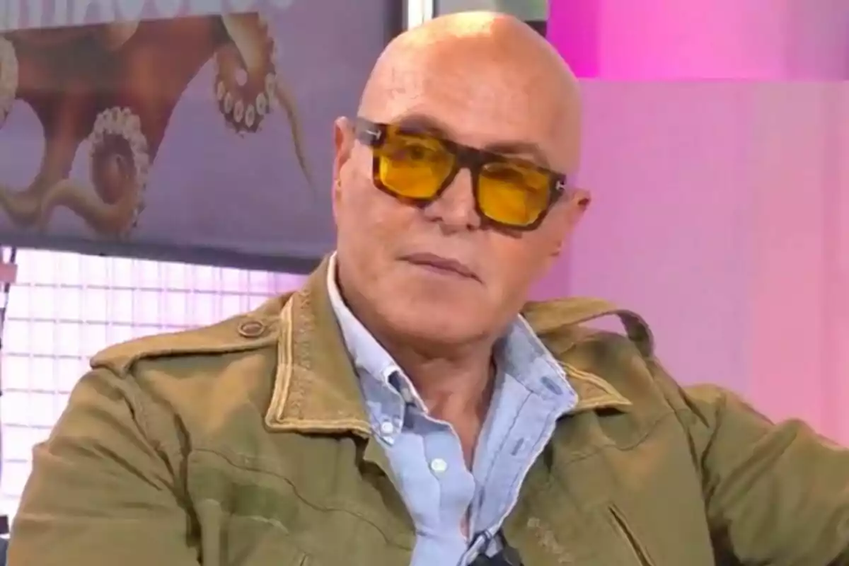 Kiko Matamoros calvo con gafas de sol amarillas y chaqueta verde está sentado en un entorno iluminado con luces rosadas y un cartel de fondo que muestra el programa 'Tentáculos'.