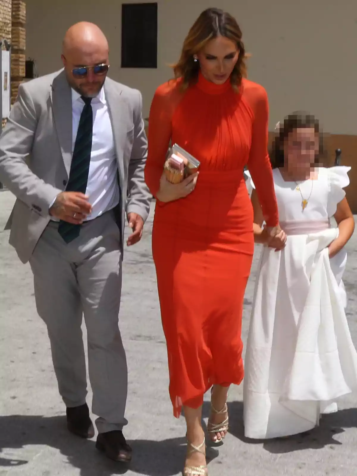 Kiko Rivera con traje gris y corbata, Irene Rosales con vestido rojo y sandalias doradas y una niña vestida de blanco caminan juntos al aire libre Kiko Rivera con traje gris y corbata, Irene Rosales con vestido rojo y sandalias doradas y una niña vestida de blanco caminan juntos al aire libre