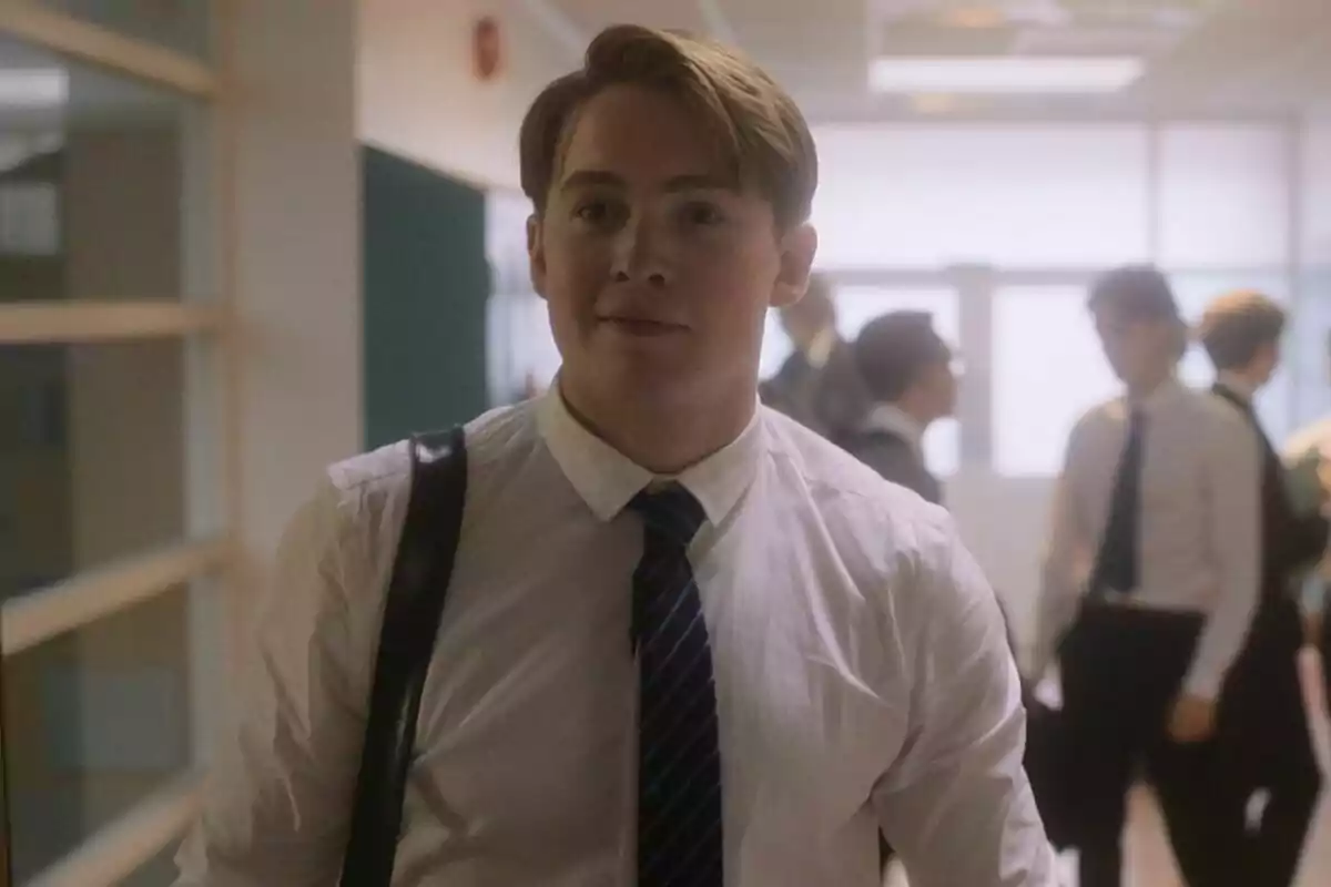 Fotografía de Kit Connor como Nick con camisa blanca y corbata azul camina por un pasillo escolar con otros estudiantes al fondo en la serie Heartstopper