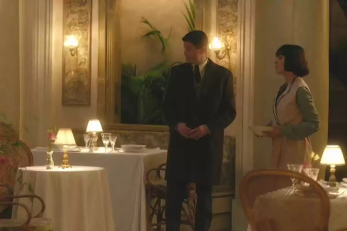 Julio y Elena está de pie en un elegante restaurante con mesas decoradas con manteles blancos, copas y lámparas de mesa, mientras conversan en un ambiente cálido y acogedor. Captura de 'La Favorita 1922' Julio y Elena está de pie en un elegante restaurante con mesas decoradas con manteles blancos, copas y lámparas de mesa, mientras conversan en un ambiente cálido y acogedor. Captura de 'La Favorita 1922'