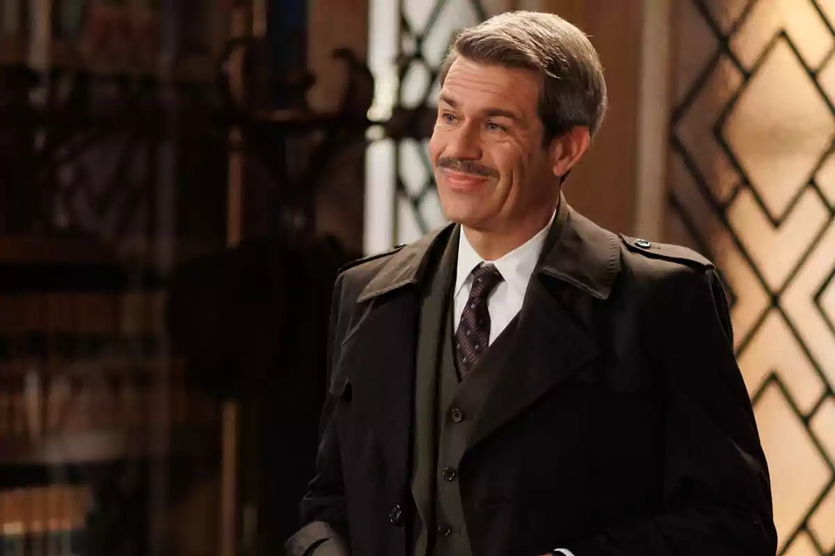 Capitán Dávila con bigote y cabello canoso, vestido con traje y abrigo, sonríe mientras está de pie en un interior elegante. Captura de 'La Moderna' Capitán Dávila con bigote y cabello canoso, vestido con traje y abrigo, sonríe mientras está de pie en un interior elegante. Captura de 'La Moderna'