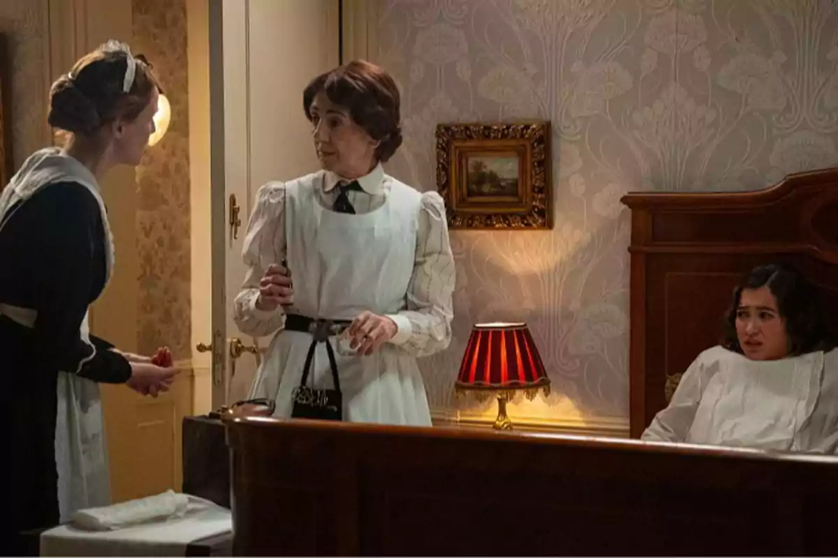 Pía, Emilia y Catalina en una habitación antigua, dos de ellas con uniforme de sirvientas y una sentada en la cama con expresión preocupada, junto a una lámpara de mesa encendida y una pared decorada con papel tapiz. Captura de 'La Promesa' Pía, Emilia y Catalina en una habitación antigua, dos de ellas con uniforme de sirvientas y una sentada en la cama con expresión preocupada, junto a una lámpara de mesa encendida y una pared decorada con papel tapiz. Captura de 'La Promesa'