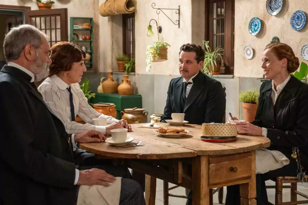Rómulo, Pía y dos personas más vestidas de época conversan sentadas alrededor de una mesa de madera con tazas y galletas en una cocina decorada con plantas y cerámica. Captura de 'La Promesa' Rómulo, Pía y dos personas más vestidas de época conversan sentadas alrededor de una mesa de madera con tazas y galletas en una cocina decorada con plantas y cerámica. Captura de 'La Promesa'