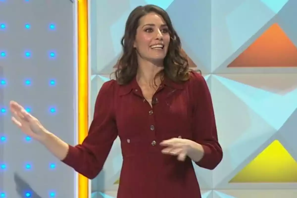 Laura Moure con vestido rojo sonríe mientras gesticula en un set de televisión con fondo geométrico y luces azules. Captura de 'La Ruleta de la Suerte'