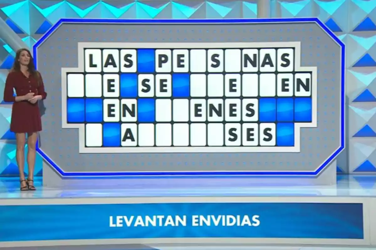 Laura Moure de pie junto a un panel de letras parcialmente resueltas en un concurso de televisión con la frase levantan envidias en la parte inferior. Captura de 'La Ruleta de la Suerte' Laura Moure de pie junto a un panel de letras parcialmente resueltas en un concurso de televisión con la frase levantan envidias en la parte inferior. Captura de 'La Ruleta de la Suerte'